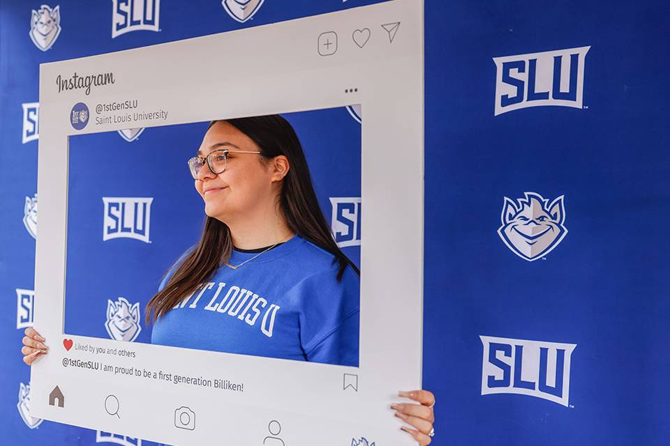 A student wearing a SLU sweatshirt stands in front of a SLU banner while holding up a picture frame that says First Generation at SLU and Proud to be a first-generation Billiken