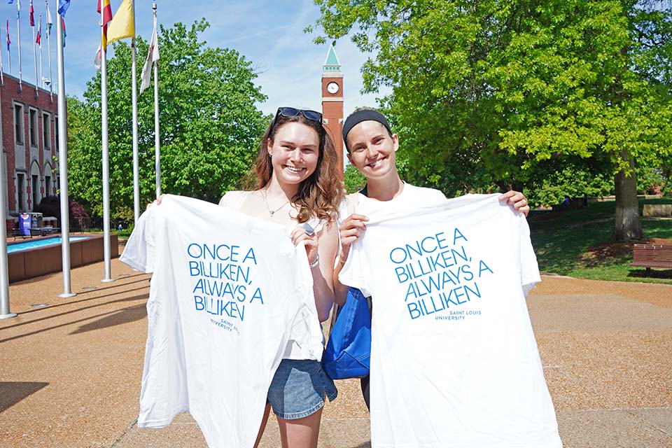 Two students pose for a photo outside on campus holding up t-shirts that sayonce a Billiken always a Billiken. 