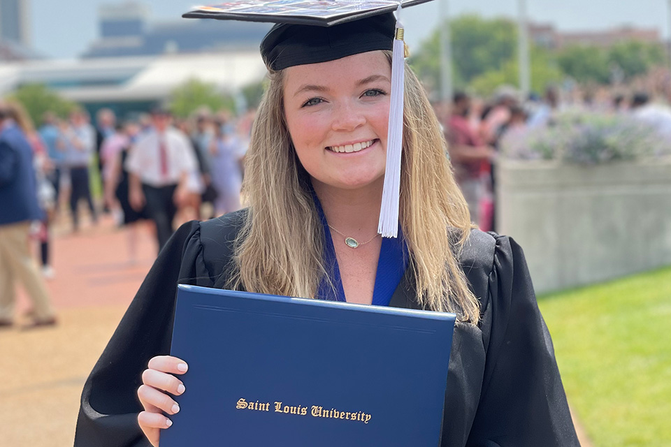 A woman wears a graduation cap and gown and holds a diploma outside on campus.