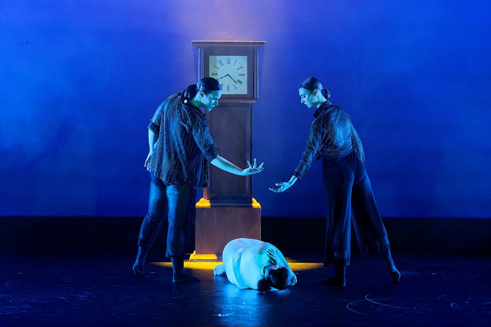 Three dancers stand in front of a clock
