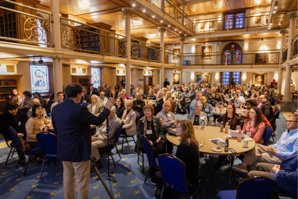 A crowded room of conference attendees are seen from the front while sitting at tables, watching a speaker