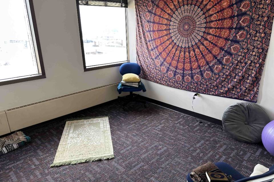 A room has a prayer rug on the floor as well as a mandala tapestry on one wall. Chairs and cushions are also visible.