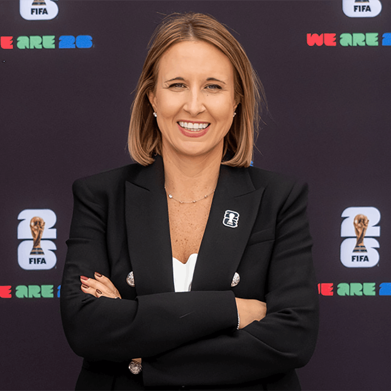 A woman in a business suit poses in front of a backdrop with the World Cup 2026 logo.