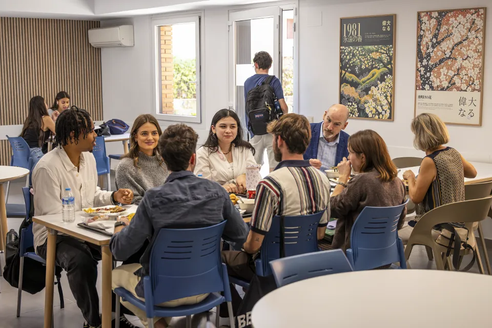 Eight people sit at a table in a cafeteria space where there are decorative posters on the wall and behind them there is a door that leads to a patio.