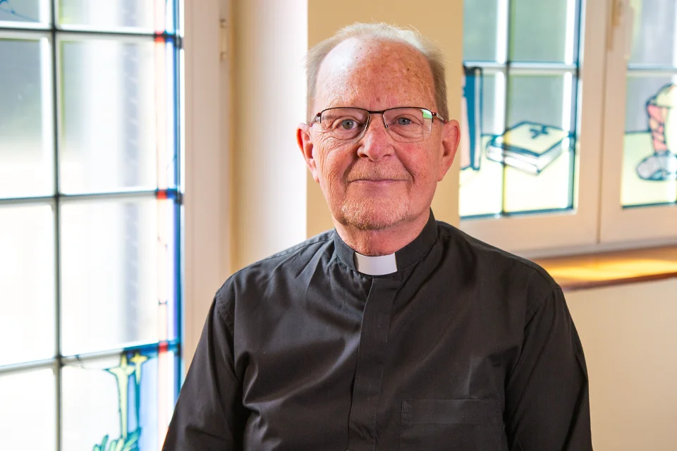 Priest poses for photo with stained glass windows in the background with religious designs.