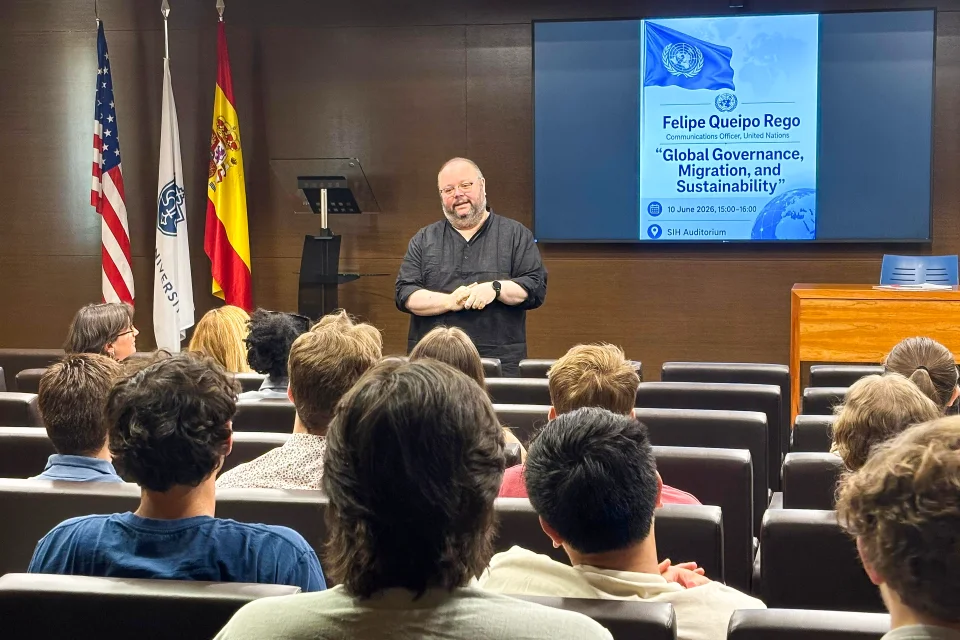 Queipo Rego speaking to students in the San Ignacio Hall Auditorium. Man speaking to students with flags of the U.S., the University, and Spain to the left and the poster of the event is projected on the screen to his right.