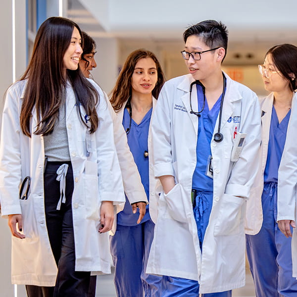 A group of medical students wearing their white coats and hospital scrubs converse while walking down a hallway.
