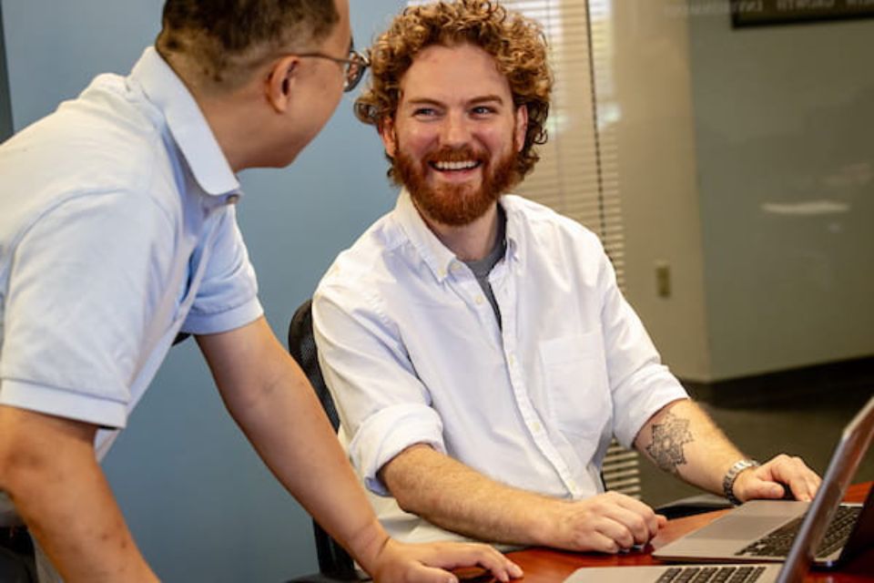 Two colleagues discussing work at a table with two computers