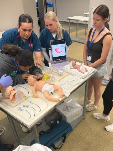 A medical instructor guides students through a simulation using an infant mannequin on a training table.