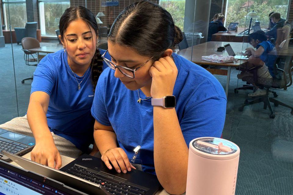 Two students look at a laptop screen together while sitting at a table in a university library.