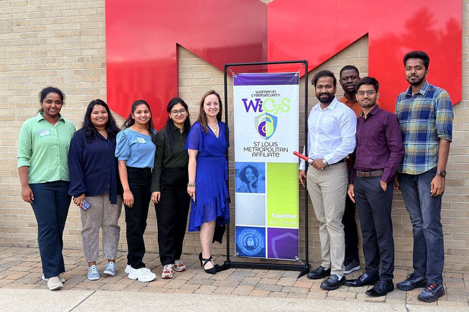 A group pose gather outdoors around a large, red "M" on the side of a building and a sign promoting an event at Maryville University.